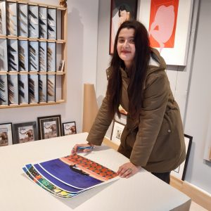 Cécile Aurégan signe ses illustrations en série limitée, imprimée d'après ses peintures originales, pour la boutique Artem Nantes. On voit son illustration "Le Chat de Tanger". Derrière l'artiste, les échantillons de baguettes pour l'encadrement sur-mesure des tableaux, une illustration de Mathilde Crétier, une sérigraphie de HelloMarine, une gravure de Cathie Pavoine, des exemples de cadres avec l'éléphant de Nantes.